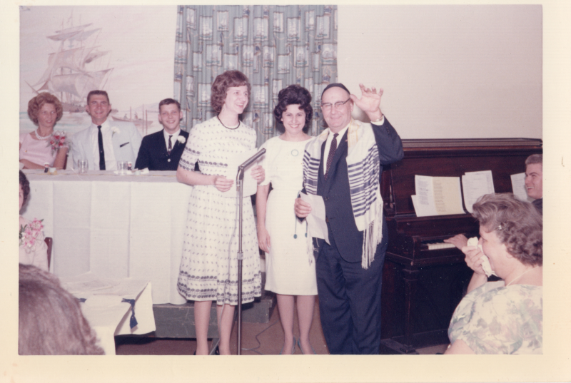 Two women and a man with a yarmulke give a speech into a microphone in front of a crowd