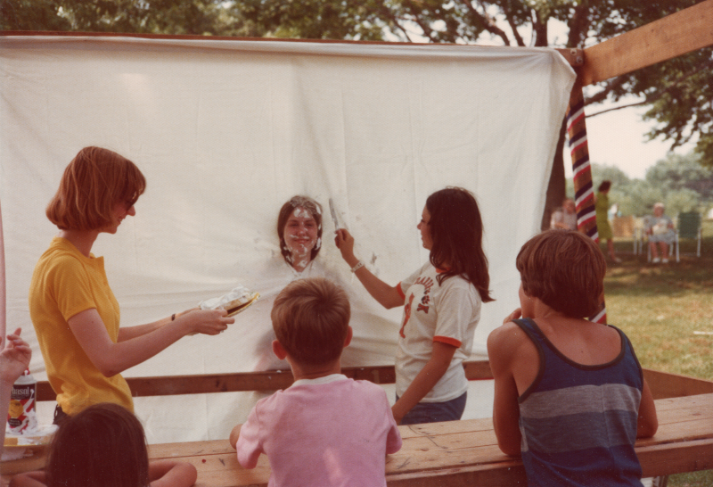 Children watch as two women pie a girl with her head peeking through a sheet of fabric