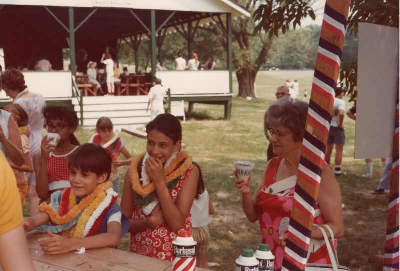 A group congregates outdoors for refreshments. Children are wearing leis. An outdoor wood structure has cans of shaving cream, and is decorated with streamers.
