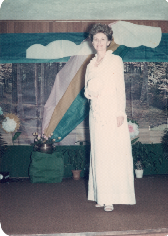 A woman in a white dress poses in front of a forestal backdrop and a rainbow