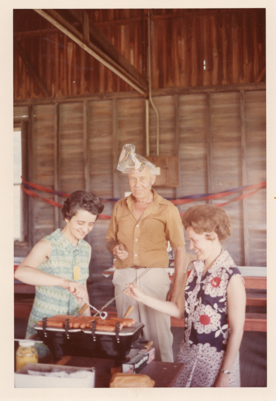 Two women cook hotdogs while a man in the background places a plastic bag on his head