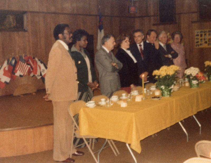 Congregants stand together in front of a table, world flags are in the background