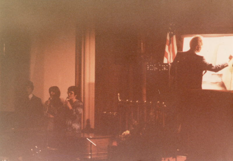 A man draws on an illuminated screen while three peeople sing