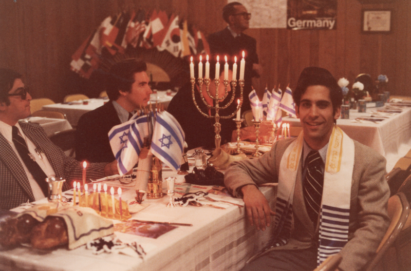 Men sit around a table with menorahs and Israeli flags. A man facing the camera waears a yarmulke and stole. There are other international flags in the background