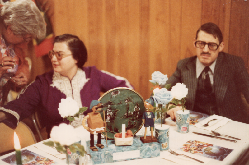 A man and women sit together at a table with small wooden figures. There are several international flags in the background