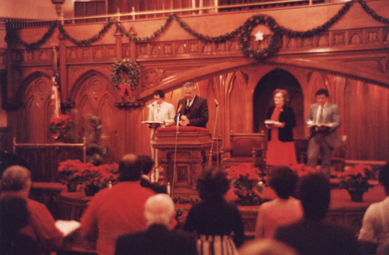 Pastor Bogdan speaks into a microphone with Mrs. Bogdan and two other men on stage in the sanctuary