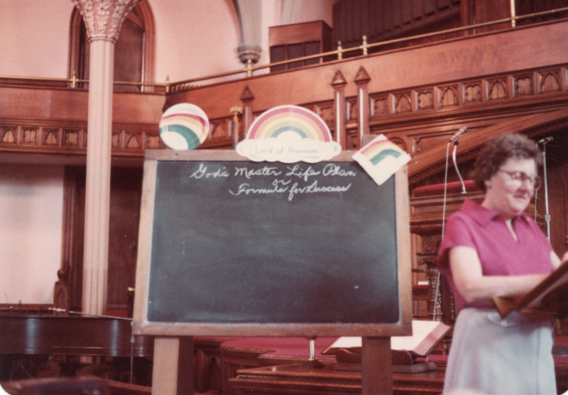 A woman in the sanctuary stands with a chalkboard decorated with ranbows reading, "God's Master Life Plan or Formula for Success"