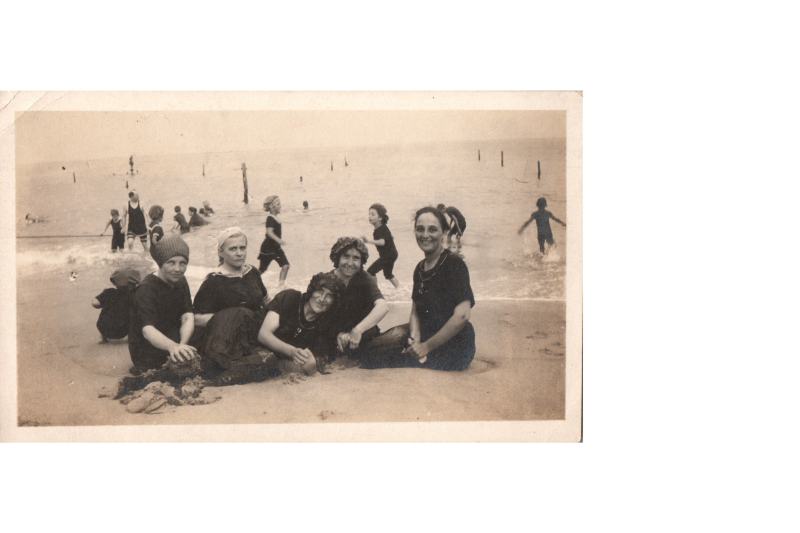 A group of women pose together at the beach with children behind them playing in the water