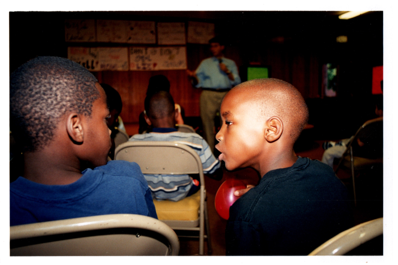 Two boys talk with Pastor Wegner speaking into a microphone in the background
