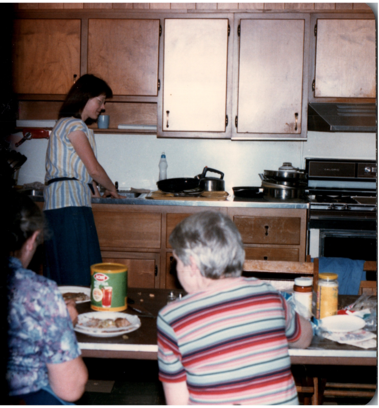 A woman prepares something in the kitchen while two women speak together